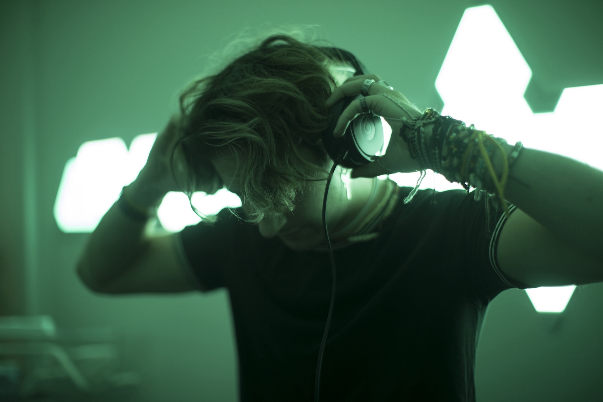 Adam-headphones-in-club-AR-DJ-Events A person with wavy hair adjusts headphones on their head in a dimly lit room with green lighting and geometric light shapes—capturing the cool, focused vibe of a corporate event DJ. Their face is partially obscured by shadows.